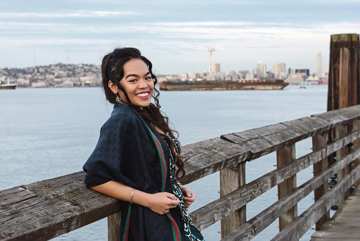 Anesha is resting back on a wood pier railing with the blue puget sound water and the Seattle skyline in the background